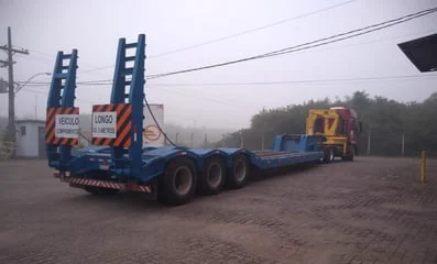 Carreta prancha rebaixada azul, sinalizada para transporte de veículo longo.